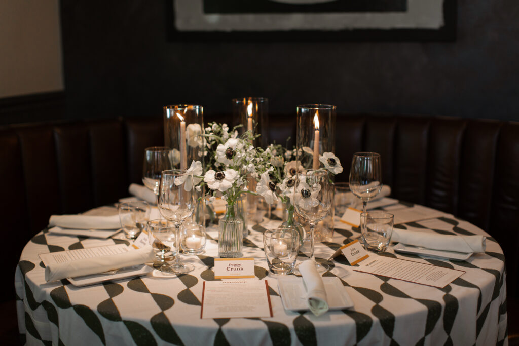 Timeless rehearsal dinner florals in a classic black and white with accents of green color palette. Bud vases and candlelight adorn the tables of this formal rehearsal dinner. Statement urn featuring olive branches and anemones decorate the bar of this reception space. Design by Rosemary & Finch Floral Design in Nashville, TN.