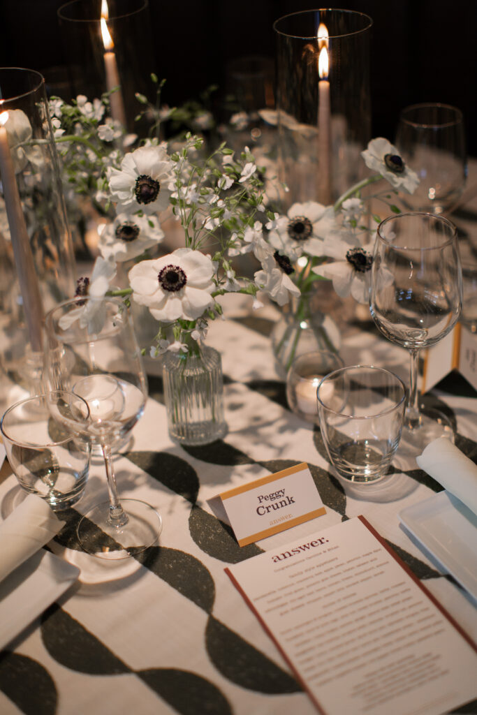 Timeless rehearsal dinner florals in a classic black and white with accents of green color palette. Bud vases and candlelight adorn the tables of this formal rehearsal dinner. Statement urn featuring olive branches and anemones decorate the bar of this reception space. Design by Rosemary & Finch Floral Design in Nashville, TN.