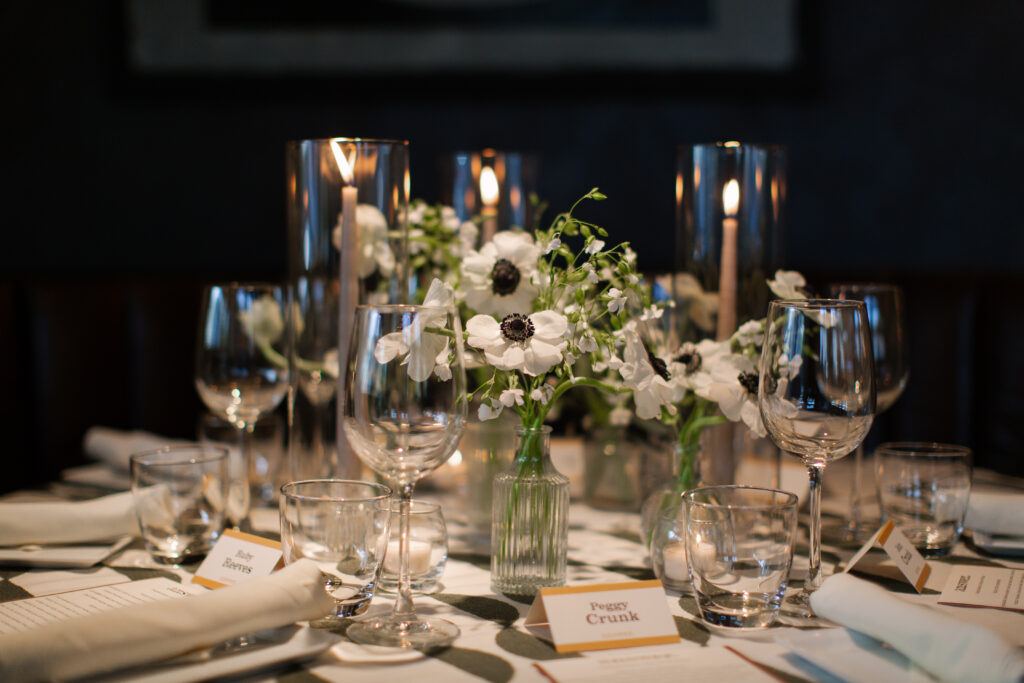 Timeless rehearsal dinner florals in a classic black and white with accents of green color palette. Bud vases and candlelight adorn the tables of this formal rehearsal dinner. Statement urn featuring olive branches and anemones decorate the bar of this reception space. Design by Rosemary & Finch Floral Design in Nashville, TN.