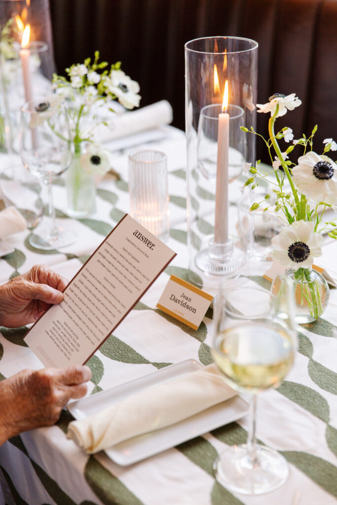 Timeless rehearsal dinner florals in a classic black and white with accents of green color palette. Bud vases and candlelight adorn the tables of this formal rehearsal dinner. Statement urn featuring olive branches and anemones decorate the bar of this reception space. Design by Rosemary & Finch Floral Design in Nashville, TN.