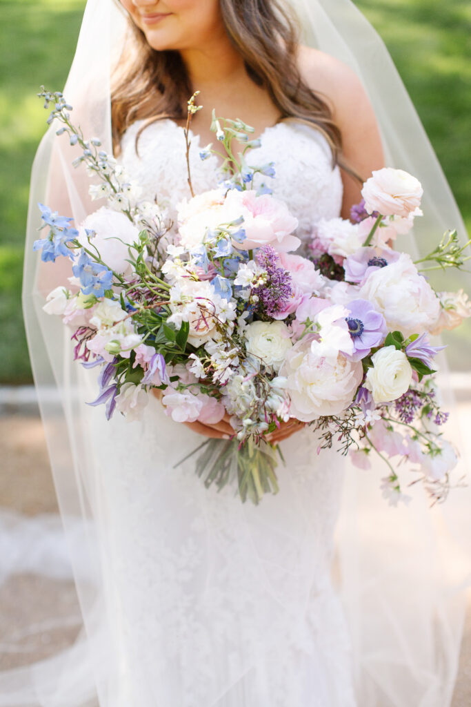 Elegant bridal bouquet in soft pastel colors, including shades of pink, white, and lavender. Mix of roses, peonies, and other delicate blooms, accented with greenery. Lush garden style bridal bouquet for spring wedding. Bridesmaids’ bouquets accenting bridal. Design by Rosemary & Finch Floral Design in Nashville, TN. 