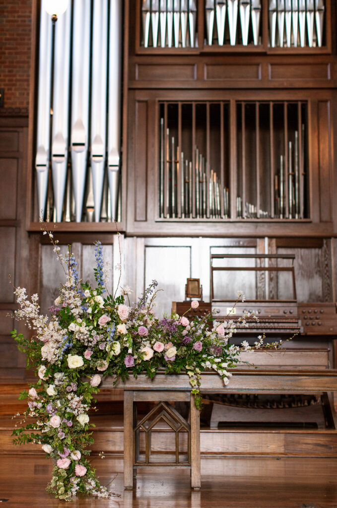 Timeless ceremony floral moments for spring wedding. Church wedding ceremony with wreath adorned doors. Lush and low floral meadows grow to accent the pews and altar of this spring wedding ceremony. Pastel color palette for spring wedding which shades of pink, purple, lavender, and cream. Mix of roses, peonies, and other delicate blooms, accented with greenery. Design by Rosemary & Finch Floral Design. 