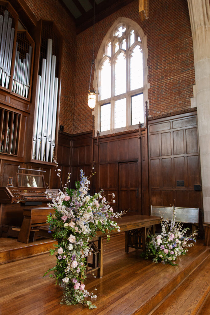 Timeless ceremony floral moments for spring wedding. Church wedding ceremony with wreath adorned doors. Lush and low floral meadows grow to accent the pews and altar of this spring wedding ceremony. Pastel color palette for spring wedding which shades of pink, purple, lavender, and cream. Mix of roses, peonies, and other delicate blooms, accented with greenery. Design by Rosemary & Finch Floral Design. 