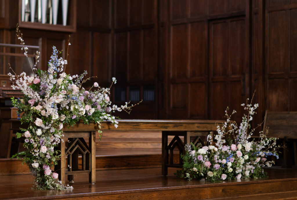 Timeless ceremony floral moments for spring wedding. Church wedding ceremony with wreath adorned doors. Lush and low floral meadows grow to accent the pews and altar of this spring wedding ceremony. Pastel color palette for spring wedding which shades of pink, purple, lavender, and cream. Mix of roses, peonies, and other delicate blooms, accented with greenery. Design by Rosemary & Finch Floral Design. 