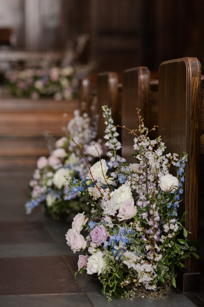 Timeless ceremony floral moments for spring wedding. Church wedding ceremony with wreath adorned doors. Lush and low floral meadows grow to accent the pews and altar of this spring wedding ceremony. Pastel color palette for spring wedding which shades of pink, purple, lavender, and cream. Mix of roses, peonies, and other delicate blooms, accented with greenery. Design by Rosemary & Finch Floral Design. 