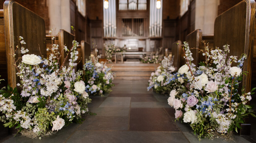 Timeless ceremony floral moments for spring wedding. Church wedding ceremony with wreath adorned doors. Lush and low floral meadows grow to accent the pews and altar of this spring wedding ceremony. Pastel color palette for spring wedding which shades of pink, purple, lavender, and cream. Mix of roses, peonies, and other delicate blooms, accented with greenery. Design by Rosemary & Finch Floral Design. 