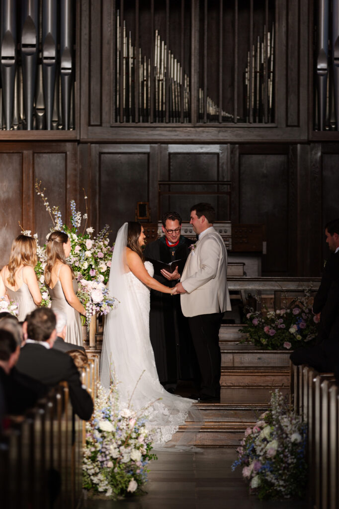 Timeless ceremony floral moments for spring wedding. Church wedding ceremony with wreath adorned doors. Lush and low floral meadows grow to accent the pews and altar of this spring wedding ceremony. Pastel color palette for spring wedding which shades of pink, purple, lavender, and cream. Mix of roses, peonies, and other delicate blooms, accented with greenery. Design by Rosemary & Finch Floral Design. 