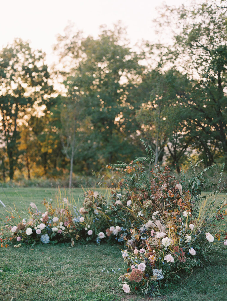 Lush garden floral meadow for fall wedding ceremony. Floral ground arch and organic aisle meadows with grassy textures and muted floral tones. Soft whimsical floral design for unique fall wedding design. Fall wedding color palette in brown, soft blue, dusty rose, and muted greens. Whimsical florals featuring dried hydrangeas, tulips, delphinium, and grasses. Design by Rosemary & Finch Floral Design in Nashville, TN. 