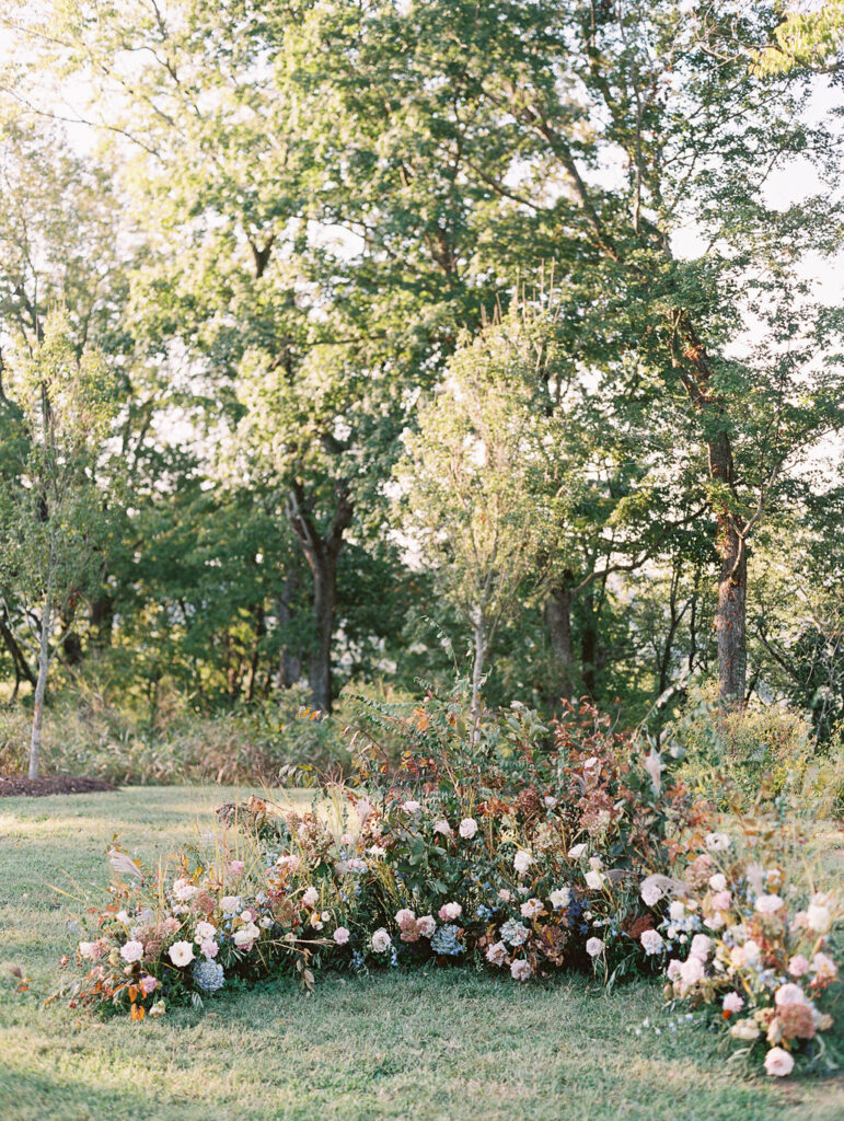 Lush garden floral meadow for fall wedding ceremony. Floral ground arch and organic aisle meadows with grassy textures and muted floral tones. Soft whimsical floral design for unique fall wedding design. Fall wedding color palette in brown, soft blue, dusty rose, and muted greens. Whimsical florals featuring dried hydrangeas, tulips, delphinium, and grasses. Design by Rosemary & Finch Floral Design in Nashville, TN. 