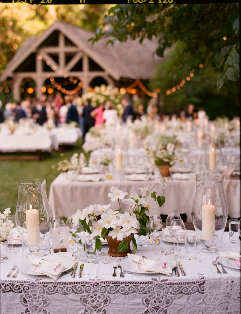 Romantic and timeless outdoor garden wedding reception featured in Vogue at Blackberry Farms. The floral design highlights white seasonal wildflowers such as cosmos, Queen Anne’s lace, sweet peas, clematis, and saponaria, with touches of texture including flowering smokebush, maidenhair fern, and herbs for a garden-inspired style atop lace table linens. Warm candlelight and soft tones create a sophisticated and intimate atmosphere. Design by Rosemary & Finch Floral Design in Nashville, TN.