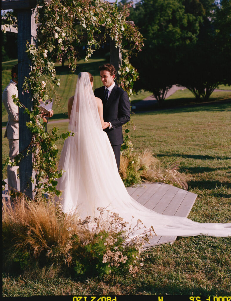 Elegant, organic and untamed outdoor wedding ceremony at Blackberry Farms featured in Vogue. Whimsical, grassy meadows line the aisle and floral accents are highlighted with white cosmos, green mist, solidago, yarrow, scabiosa and grasses. Pergola is covered in lush greenery with soft touches of cream roses and wispy meadows at the base. Design by Rosemary & Finch Floral Design in Nashville, TN. 