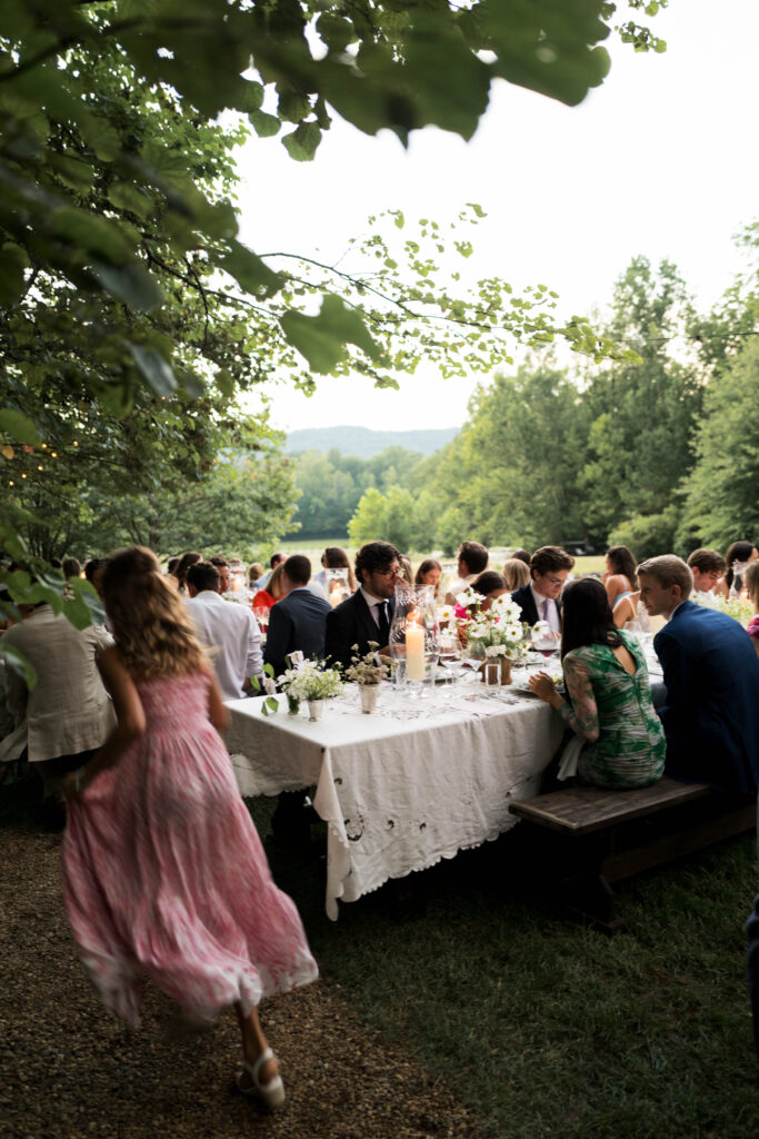 Romantic and timeless outdoor garden wedding reception featured in Vogue at Blackberry Farms. The floral design highlights white seasonal wildflowers such as cosmos, Queen Anne’s lace, sweet peas, clematis, and saponaria, with touches of texture including flowering smokebush, maidenhair fern, and herbs for a garden-inspired style atop lace table linens. Warm candlelight and soft tones create a sophisticated and intimate atmosphere. Design by Rosemary & Finch Floral Design in Nashville, TN.
