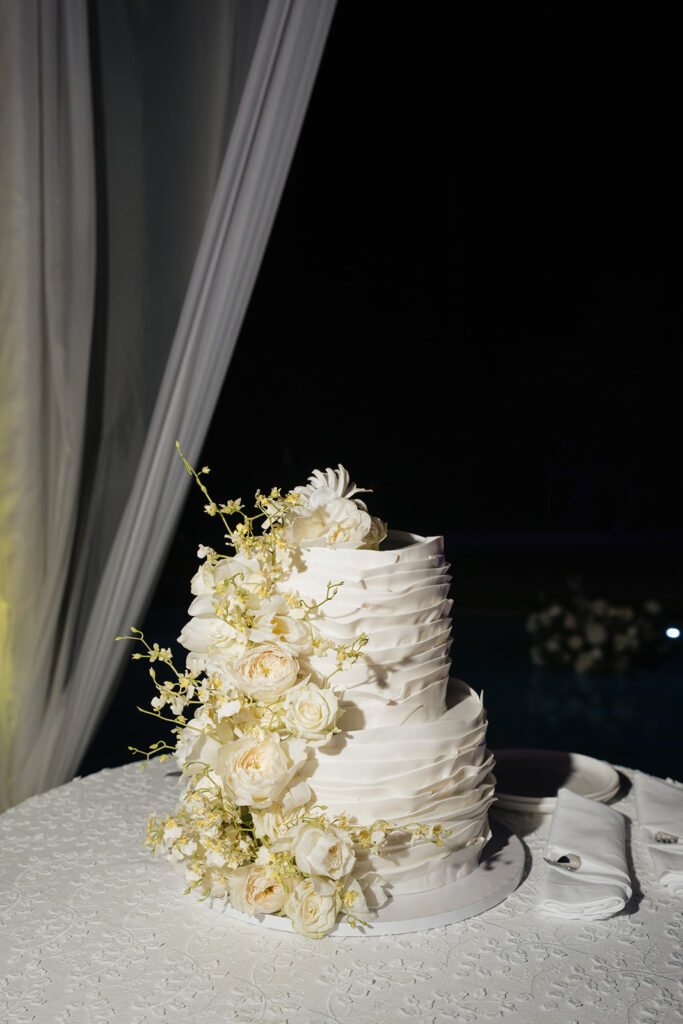Luxurious, all-white flowers cascading over a stunning wedding cake. Flowers featured are roses, ranunculus, oncidium, delphinium, spray roses and waxflower with a slight amount of greenery. The cake is surrounded by all-white, lush meadows placed on the floor. Meadows include cake flowers as well as lisianthus, hydrangeas, baby’s breath, tulips, mums and more. Design by Rosemary & Finch Floral Design in Nashville, TN. 