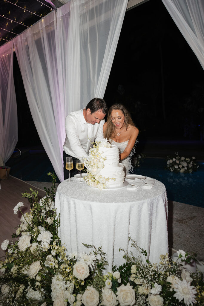 Luxurious, all-white flowers cascading over a stunning wedding cake. Flowers featured are roses, ranunculus, oncidium, delphinium, spray roses and waxflower with a slight amount of greenery. The cake is surrounded by all-white, lush meadows placed on the floor. Meadows include cake flowers as well as lisianthus, hydrangeas, baby’s breath, tulips, mums and more. Design by Rosemary & Finch Floral Design in Nashville, TN. 