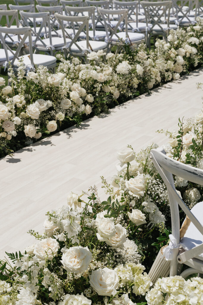 Lush white ceremony florals for a beachfront wedding in the Bahamas featuring garden roses, hydrangeas, delphinium, spray roses, baby’s breath, chrysanthemums, waxflower, carnations, tulips and a mixture of greens to create a refined and romantic aisle arrangement. Layered textures and soft movement give these all-white flowers an elevated, editorial look. Design by Rosemary & Finch Floral Design in Nashville, TN.