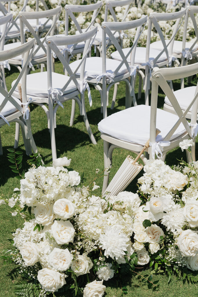 Lush white ceremony florals for a beachfront wedding in the Bahamas featuring garden roses, hydrangeas, delphinium, spray roses, baby’s breath, chrysanthemums, waxflower, carnations, tulips and a mixture of greens to create a refined and romantic aisle arrangement. Layered textures and soft movement give these all-white flowers an elevated, editorial look. Design by Rosemary & Finch Floral Design in Nashville, TN.