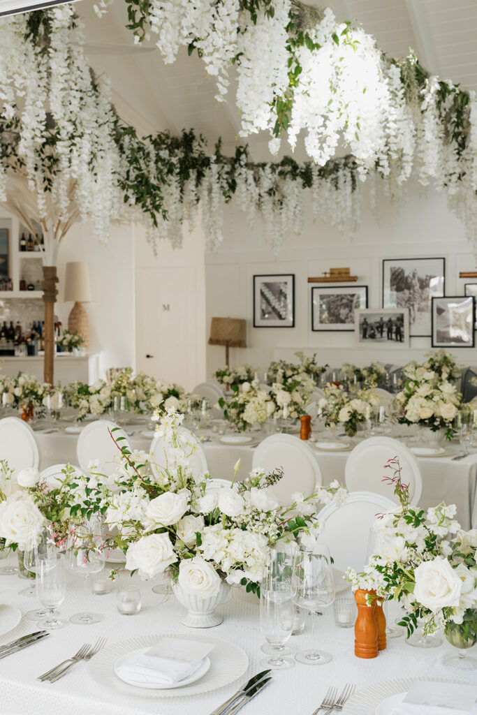 Ethereal white wedding reception with a breathtaking canopy of cascading white wisteria & green vines. Tables feature low, lush centerpieces combining garden roses, hydrangeas, spray roses, waxflower, delphinium, ranunculus, tulips, oncidium orchids, lisianthus, tweedia, lilac, sweet peas and are accented by deep-green foliage. This stunning floral design creates a luxurious, romantic, all-white atmosphere. Design by Rosemary & Finch Floral Design in Nashville, TN. 