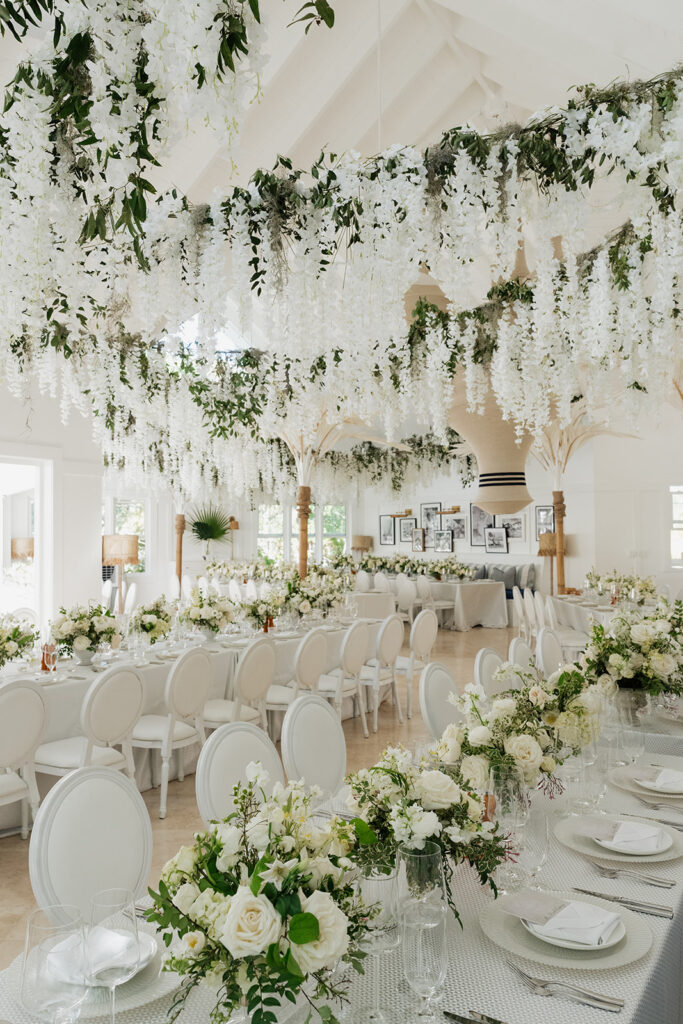 Ethereal white wedding reception with a breathtaking canopy of cascading white wisteria & green vines. Tables feature low, lush centerpieces combining garden roses, hydrangeas, spray roses, waxflower, delphinium, ranunculus, tulips, oncidium orchids, lisianthus, tweedia, lilac, sweet peas and are accented by deep-green foliage. This stunning floral design creates a luxurious, romantic, all-white atmosphere. Design by Rosemary & Finch Floral Design in Nashville, TN. 