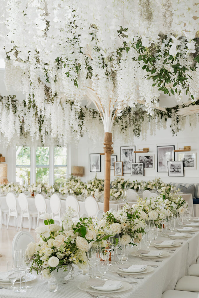 Ethereal white wedding reception with a breathtaking canopy of cascading white wisteria & green vines. Tables feature low, lush centerpieces combining garden roses, hydrangeas, spray roses, waxflower, delphinium, ranunculus, tulips, oncidium orchids, lisianthus, tweedia, lilac, sweet peas and are accented by deep-green foliage. This stunning floral design creates a luxurious, romantic, all-white atmosphere. Design by Rosemary & Finch Floral Design in Nashville, TN. 