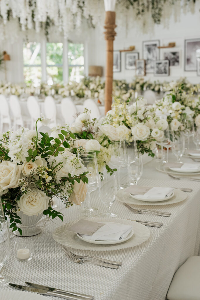 Ethereal white wedding reception with a breathtaking canopy of cascading white wisteria & green vines. Tables feature low, lush centerpieces combining garden roses, hydrangeas, spray roses, waxflower, delphinium, ranunculus, tulips, oncidium orchids, lisianthus, tweedia, lilac, sweet peas and are accented by deep-green foliage. This stunning floral design creates a luxurious, romantic, all-white atmosphere. Design by Rosemary & Finch Floral Design in Nashville, TN. 