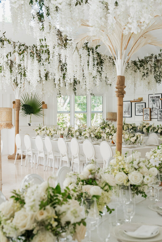Ethereal white wedding reception with a breathtaking canopy of cascading white wisteria & green vines. Tables feature low, lush centerpieces combining garden roses, hydrangeas, spray roses, waxflower, delphinium, ranunculus, tulips, oncidium orchids, lisianthus, tweedia, lilac, sweet peas and are accented by deep-green foliage. This stunning floral design creates a luxurious, romantic, all-white atmosphere. Design by Rosemary & Finch Floral Design in Nashville, TN. 