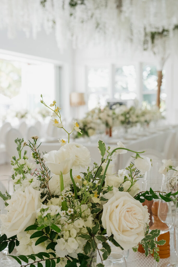 Ethereal white wedding reception with a breathtaking canopy of cascading white wisteria & green vines. Tables feature low, lush centerpieces combining garden roses, hydrangeas, spray roses, waxflower, delphinium, ranunculus, tulips, oncidium orchids, lisianthus, tweedia, lilac, sweet peas and are accented by deep-green foliage. This stunning floral design creates a luxurious, romantic, all-white atmosphere. Design by Rosemary & Finch Floral Design in Nashville, TN. 
