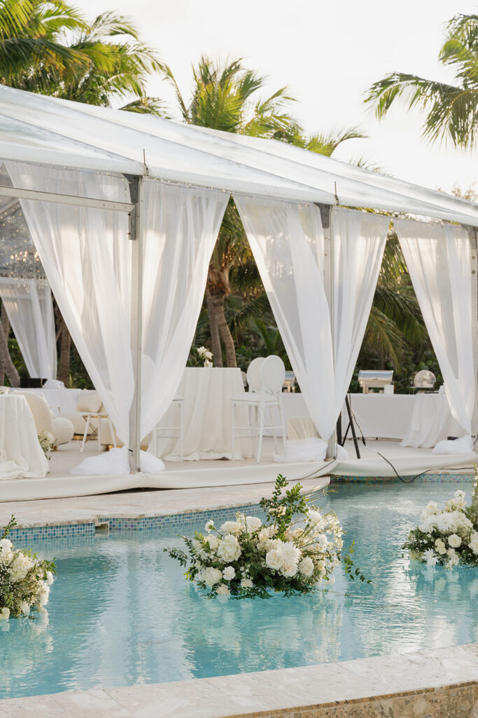 Elegant floating wedding florals in a gorgeous pool with a palm tree backdrop in the Bahamas. The arrangements feature abundant white hydrangeas, spray roses, garden cloud roses, waxflower, delphinium, ranunculus, tulips, oncidium orchids, lisianthus, tweedia, lilac, sweet peas and more. Luxurious all-white event decor for a destination wedding in the Bahamas. Design by Rosemary & Finch Floral Design in Nashville, TN. 
