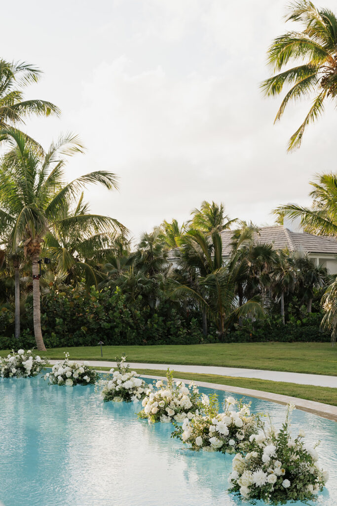 Elegant floating wedding florals in a gorgeous pool with a palm tree backdrop in the Bahamas. The arrangements feature abundant white hydrangeas, spray roses, garden cloud roses, waxflower, delphinium, ranunculus, tulips, oncidium orchids, lisianthus, tweedia, lilac, sweet peas and more. Luxurious all-white event decor for a destination wedding in the Bahamas. Design by Rosemary & Finch Floral Design in Nashville, TN. 
