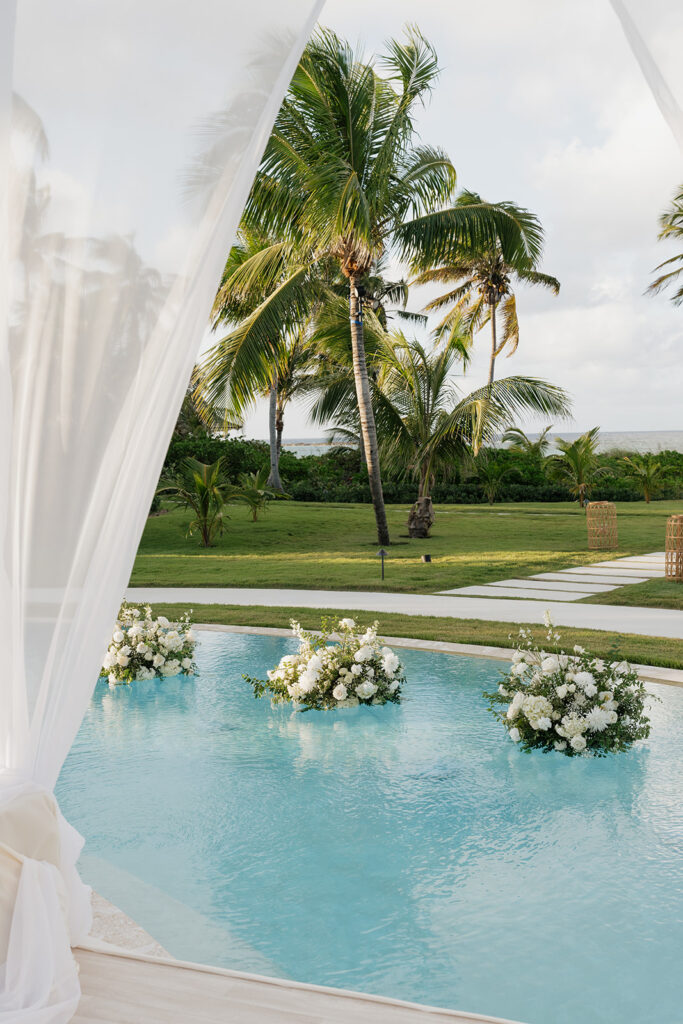 Elegant floating wedding florals in a gorgeous pool with a palm tree backdrop in the Bahamas. The arrangements feature abundant white hydrangeas, spray roses, garden cloud roses, waxflower, delphinium, ranunculus, tulips, oncidium orchids, lisianthus, tweedia, lilac, sweet peas and more. Luxurious all-white event decor for a destination wedding in the Bahamas. Design by Rosemary & Finch Floral Design in Nashville, TN. 
