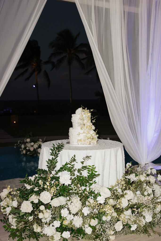 Luxurious, all-white flowers cascading over a stunning wedding cake. Flowers featured are roses, ranunculus, oncidium, delphinium, spray roses and waxflower with a slight amount of greenery. The cake is surrounded by all-white, lush meadows placed on the floor. Meadows include cake flowers as well as lisianthus, hydrangeas, baby’s breath, tulips, mums and more. Design by Rosemary & Finch Floral Design in Nashville, TN. 
