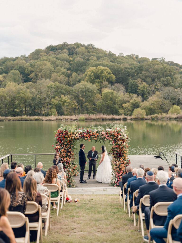 Stunning flower-forward, lush chuppah for fall wedding ceremony at Southall Farm and Inn. Bright, autumnal colors of rosy pink, deep red, blush, rusty orange, peach, and golden yellow are combined with the depth of full greenery. Color-blocked florals of dahlias, tess roses, cappuccino roses, moab roses, quicksand roses, spray roses, zinnias, lisianthus, yarrow, marigolds, ranunculus, and more cover the chuppah. Design by Rosemary and Finch Floral Design in Nashville, TN.