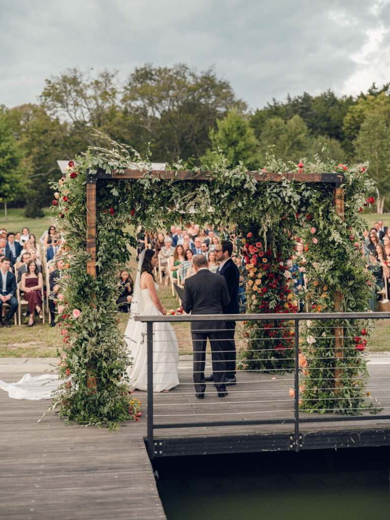 Stunning flower-forward, lush chuppah for fall wedding ceremony at Southall Farm and Inn. Bright, autumnal colors of rosy pink, deep red, blush, rusty orange, peach, and golden yellow are combined with the depth of full greenery. Color-blocked florals of dahlias, tess roses, cappuccino roses, moab roses, quicksand roses, spray roses, zinnias, lisianthus, yarrow, marigolds, ranunculus, and more cover the chuppah. Design by Rosemary and Finch Floral Design in Nashville, TN.