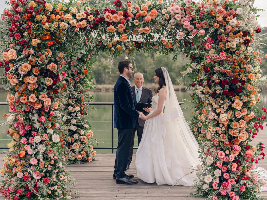Stunning flower-forward, lush chuppah for fall wedding ceremony at Southall Farm and Inn. Bright, autumnal colors of rosy pink, deep red, blush, rusty orange, peach, and golden yellow are combined with the depth of full greenery. Color-blocked florals of dahlias, tess roses, cappuccino roses, moab roses, quicksand roses, spray roses, zinnias, lisianthus, yarrow, marigolds, ranunculus, and more cover the chuppah. Design by Rosemary and Finch Floral Design in Nashville, TN.