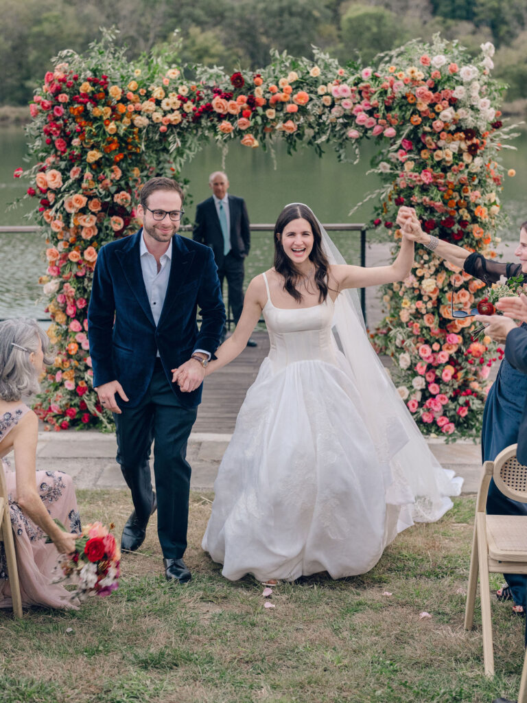 Stunning flower-forward, lush chuppah for fall wedding ceremony at Southall Farm and Inn. Bright, autumnal colors of rosy pink, deep red, blush, rusty orange, peach, and golden yellow are combined with the depth of full greenery. Color-blocked florals of dahlias, tess roses, cappuccino roses, moab roses, quicksand roses, spray roses, zinnias, lisianthus, yarrow, marigolds, ranunculus, and more cover the chuppah. Design by Rosemary and Finch Floral Design in Nashville, TN.