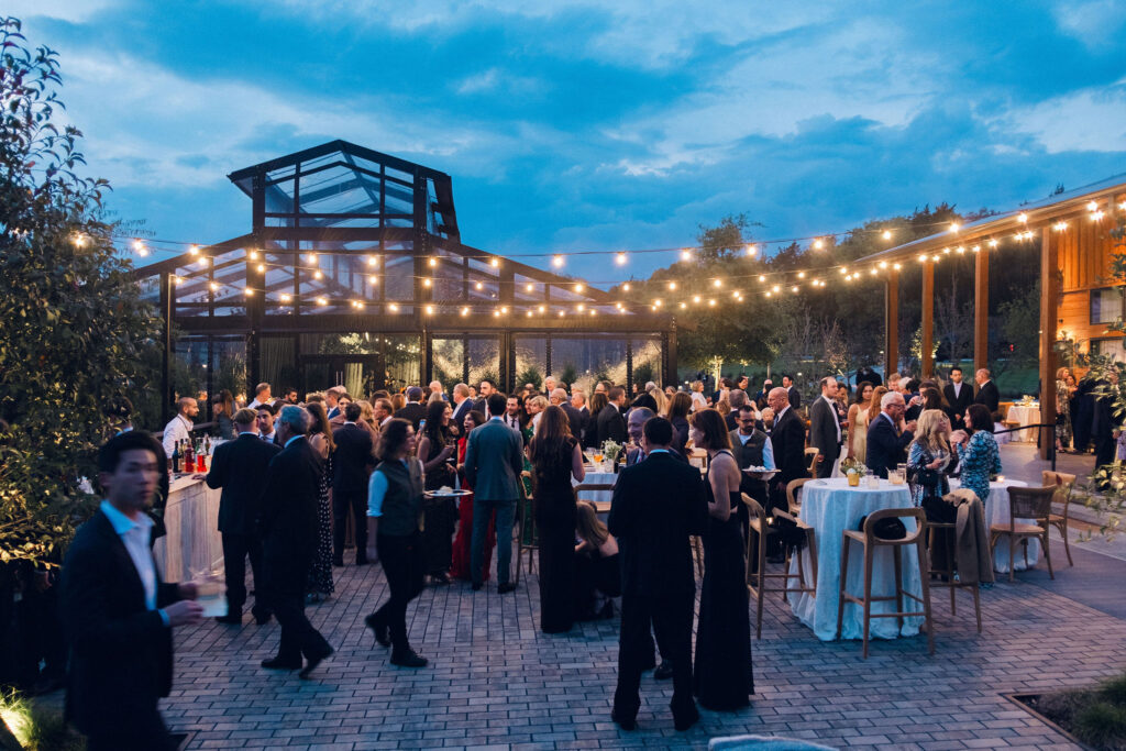 Elegant outdoor cocktail hour with bud vase floral arrangements scattered throughout high boy tables. All-white roses, lisianthus, delphinium, spray roses, ranunculus, campunala, saponaria, and light greenery were used for bud vases to correspond with floral reception centerpieces. The escort display was highlighted with chamomile, flannel flower, ranunculus, cloud garden roses, spray roses and greenery. Design by Rosemary and Finch Floral Design in Nashville, TN. 