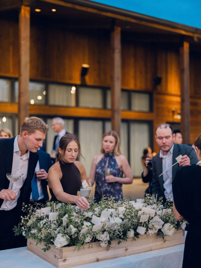 Elegant outdoor cocktail hour with bud vase floral arrangements scattered throughout high boy tables. All-white roses, lisianthus, delphinium, spray roses, ranunculus, campunala, saponaria, and light greenery were used for bud vases to correspond with floral reception centerpieces. The escort display was highlighted with chamomile, flannel flower, ranunculus, cloud garden roses, spray roses and greenery. Design by Rosemary and Finch Floral Design in Nashville, TN. 