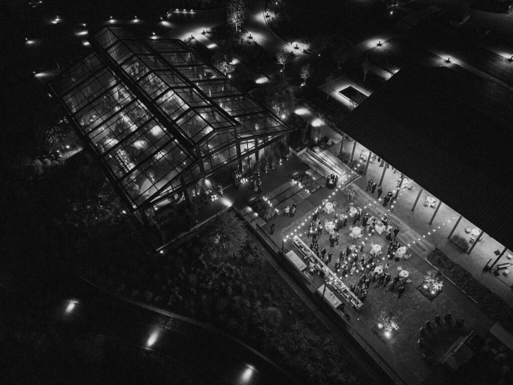 A moody, garden-inspired wedding reception featuring elegant white and green floral centerpieces under a glass tent for fall wedding. Cloud roses, ranunculus, lisianthus, chamomile, queen anne’s lace, saponaria, delphinium, flannel flower and delicate greenery create a heavy floral focal point for tables in tones of white, cream and green with amber table accents and warm candle lighting. Lush greenery drapes across the ceiling. Design by Rosemary and Finch Floral Design in Nashville, TN. 
