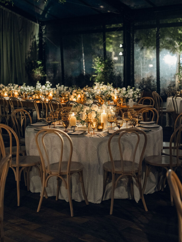 A moody, garden-inspired wedding reception featuring elegant white and green floral centerpieces under a glass tent for fall wedding. Cloud roses, ranunculus, lisianthus, chamomile, queen anne’s lace, saponaria, delphinium, flannel flower and delicate greenery create a heavy floral focal point for tables in tones of white, cream and green with amber table accents and warm candle lighting. Lush greenery drapes across the ceiling. Design by Rosemary and Finch Floral Design in Nashville, TN. 