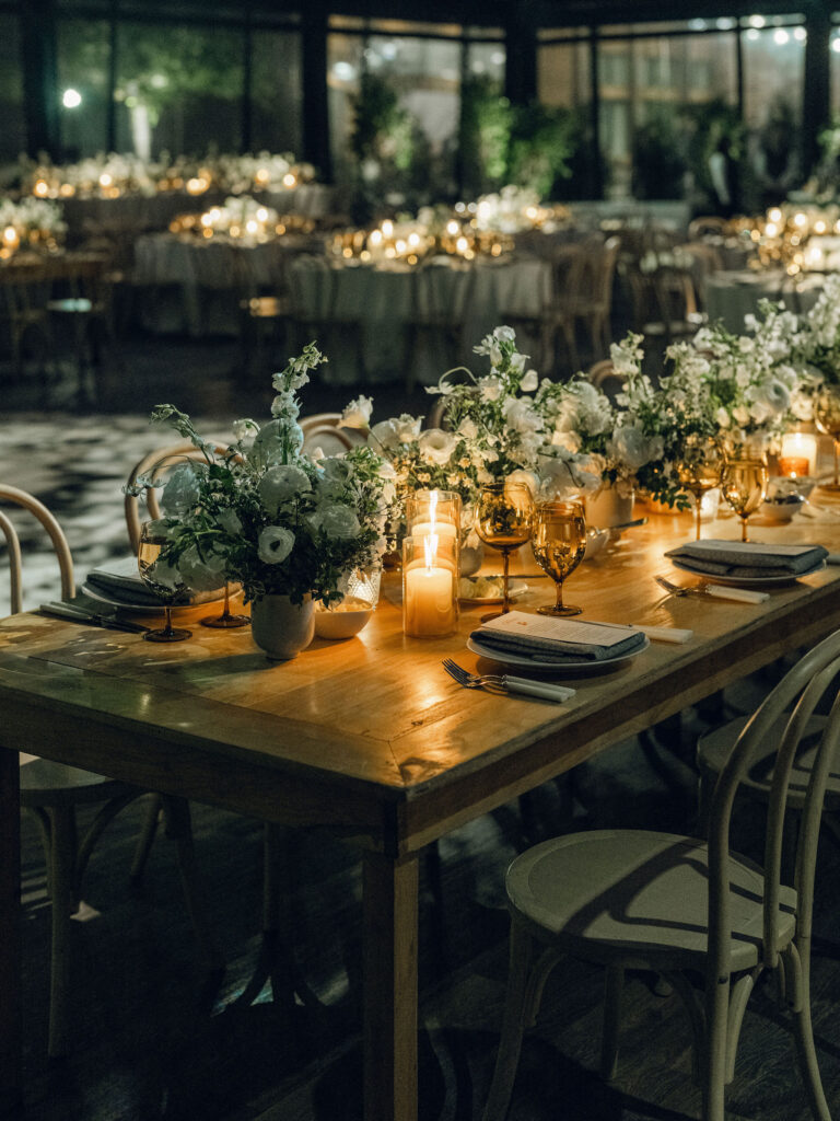 A moody, garden-inspired wedding reception featuring elegant white and green floral centerpieces under a glass tent for fall wedding. Cloud roses, ranunculus, lisianthus, chamomile, queen anne’s lace, saponaria, delphinium, flannel flower and delicate greenery create a heavy floral focal point for tables in tones of white, cream and green with amber table accents and warm candle lighting. Lush greenery drapes across the ceiling. Design by Rosemary and Finch Floral Design in Nashville, TN. 