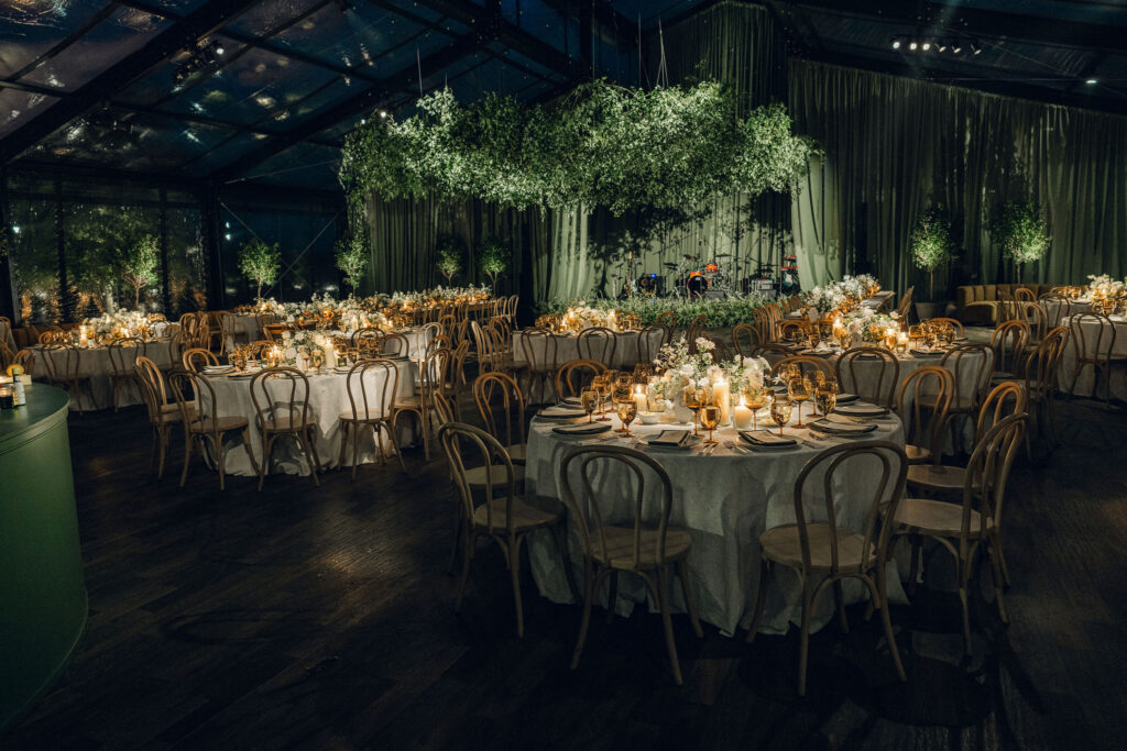 A moody, garden-inspired wedding reception featuring elegant white and green floral centerpieces under a glass tent for fall wedding. Cloud roses, ranunculus, lisianthus, chamomile, queen anne’s lace, saponaria, delphinium, flannel flower and delicate greenery create a heavy floral focal point for tables in tones of white, cream and green with amber table accents and warm candle lighting. Lush greenery drapes across the ceiling. Design by Rosemary and Finch Floral Design in Nashville, TN. 