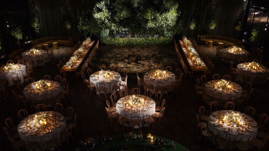 A moody, garden-inspired wedding reception featuring elegant white and green floral centerpieces under a glass tent for fall wedding. Cloud roses, ranunculus, lisianthus, chamomile, queen anne’s lace, saponaria, delphinium, flannel flower and delicate greenery create a heavy floral focal point for tables in tones of white, cream and green with amber table accents and warm candle lighting. Lush greenery drapes across the ceiling. Design by Rosemary and Finch Floral Design in Nashville, TN. 