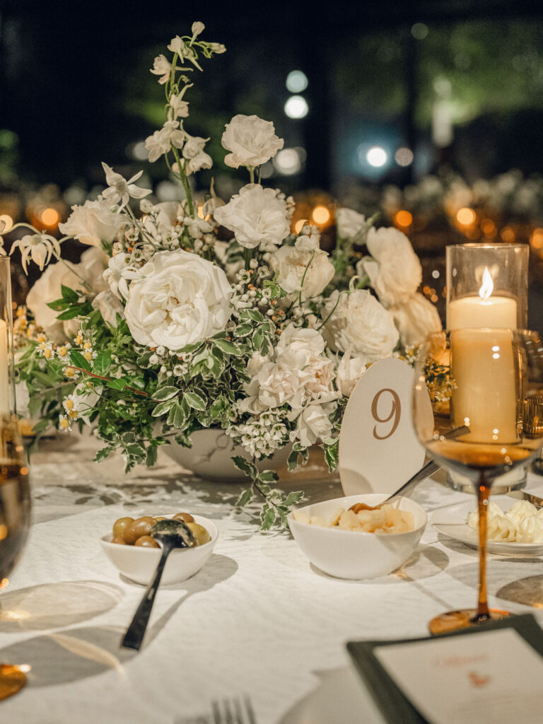 A moody, garden-inspired wedding reception featuring elegant white and green floral centerpieces under a glass tent for fall wedding. Cloud roses, ranunculus, lisianthus, chamomile, queen anne’s lace, saponaria, delphinium, flannel flower and delicate greenery create a heavy floral focal point for tables in tones of white, cream and green with amber table accents and warm candle lighting. Lush greenery drapes across the ceiling. Design by Rosemary and Finch Floral Design in Nashville, TN. 