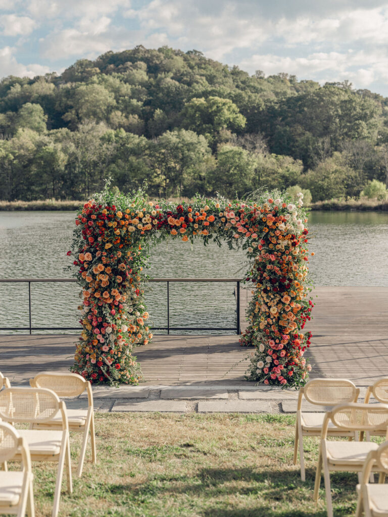 Stunning flower-forward, lush chuppah for fall wedding ceremony at Southall Farm and Inn. Bright, autumnal colors of rosy pink, deep red, blush, rusty orange, peach, and golden yellow are combined with the depth of full greenery. Color-blocked florals of dahlias, tess roses, cappuccino roses, moab roses, quicksand roses, spray roses, zinnias, lisianthus, yarrow, marigolds, ranunculus, and more cover the chuppah. Design by Rosemary and Finch Floral Design in Nashville, TN.