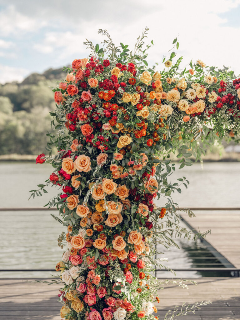 Stunning flower-forward, lush chuppah for fall wedding ceremony at Southall Farm and Inn. Bright, autumnal colors of rosy pink, deep red, blush, rusty orange, peach, and golden yellow are combined with the depth of full greenery. Color-blocked florals of dahlias, tess roses, cappuccino roses, moab roses, quicksand roses, spray roses, zinnias, lisianthus, yarrow, marigolds, ranunculus, and more cover the chuppah. Design by Rosemary and Finch Floral Design in Nashville, TN.