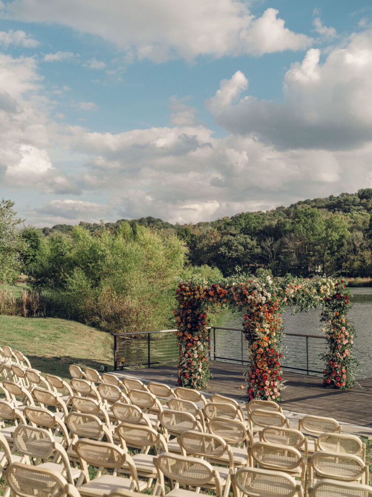 Stunning flower-forward, lush chuppah for fall wedding ceremony at Southall Farm and Inn. Bright, autumnal colors of rosy pink, deep red, blush, rusty orange, peach, and golden yellow are combined with the depth of full greenery. Color-blocked florals of dahlias, tess roses, cappuccino roses, moab roses, quicksand roses, spray roses, zinnias, lisianthus, yarrow, marigolds, ranunculus, and more cover the chuppah. Design by Rosemary and Finch Floral Design in Nashville, TN.