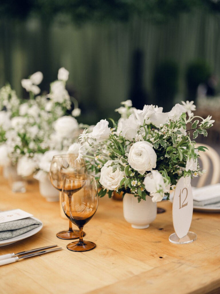 A moody, garden-inspired wedding reception featuring elegant white and green floral centerpieces under a glass tent for fall wedding. Cloud roses, ranunculus, lisianthus, chamomile, queen anne’s lace, saponaria, delphinium, flannel flower and delicate greenery create a heavy floral focal point for tables in tones of white, cream and green with amber table accents and warm candle lighting. Lush greenery drapes across the ceiling. Design by Rosemary and Finch Floral Design in Nashville, TN. 