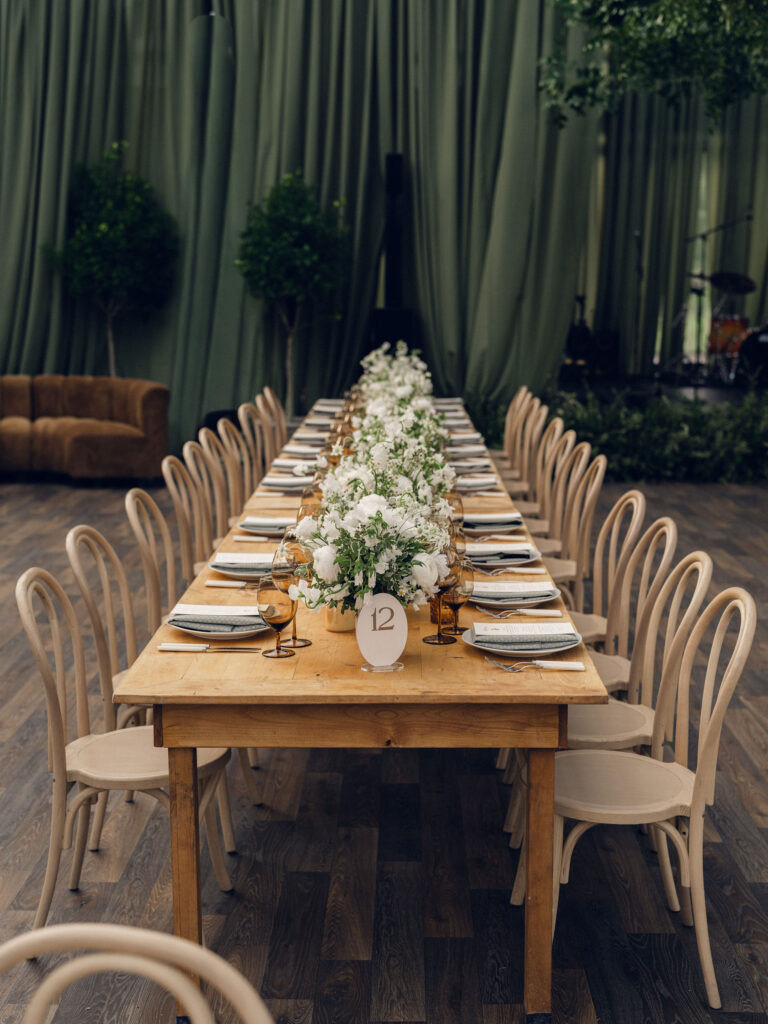 A moody, garden-inspired wedding reception featuring elegant white and green floral centerpieces under a glass tent for fall wedding. Cloud roses, ranunculus, lisianthus, chamomile, queen anne’s lace, saponaria, delphinium, flannel flower and delicate greenery create a heavy floral focal point for tables in tones of white, cream and green with amber table accents and warm candle lighting. Lush greenery drapes across the ceiling. Design by Rosemary and Finch Floral Design in Nashville, TN. 