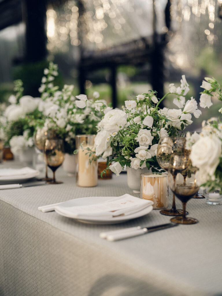 A moody, garden-inspired wedding reception featuring elegant white and green floral centerpieces under a glass tent for fall wedding. Cloud roses, ranunculus, lisianthus, chamomile, queen anne’s lace, saponaria, delphinium, flannel flower and delicate greenery create a heavy floral focal point for tables in tones of white, cream and green with amber table accents and warm candle lighting. Lush greenery drapes across the ceiling. Design by Rosemary and Finch Floral Design in Nashville, TN. 