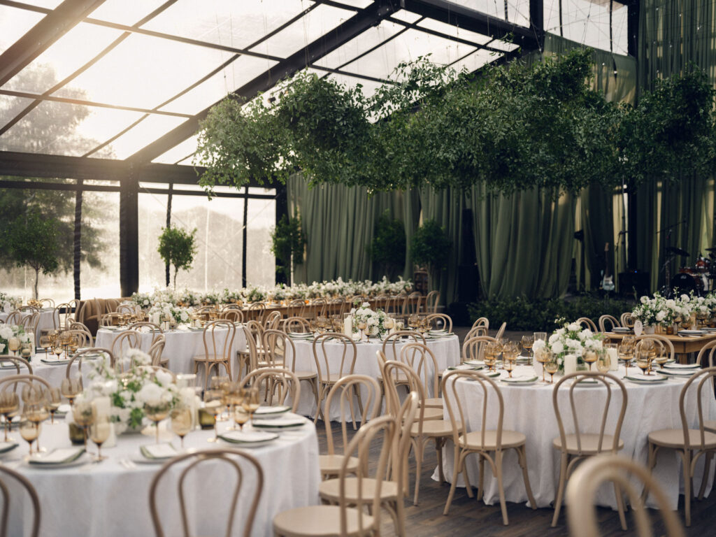 A moody, garden-inspired wedding reception featuring elegant white and green floral centerpieces under a glass tent for fall wedding. Cloud roses, ranunculus, lisianthus, chamomile, queen anne’s lace, saponaria, delphinium, flannel flower and delicate greenery create a heavy floral focal point for tables in tones of white, cream and green with amber table accents and warm candle lighting. Lush greenery drapes across the ceiling. Design by Rosemary and Finch Floral Design in Nashville, TN. 