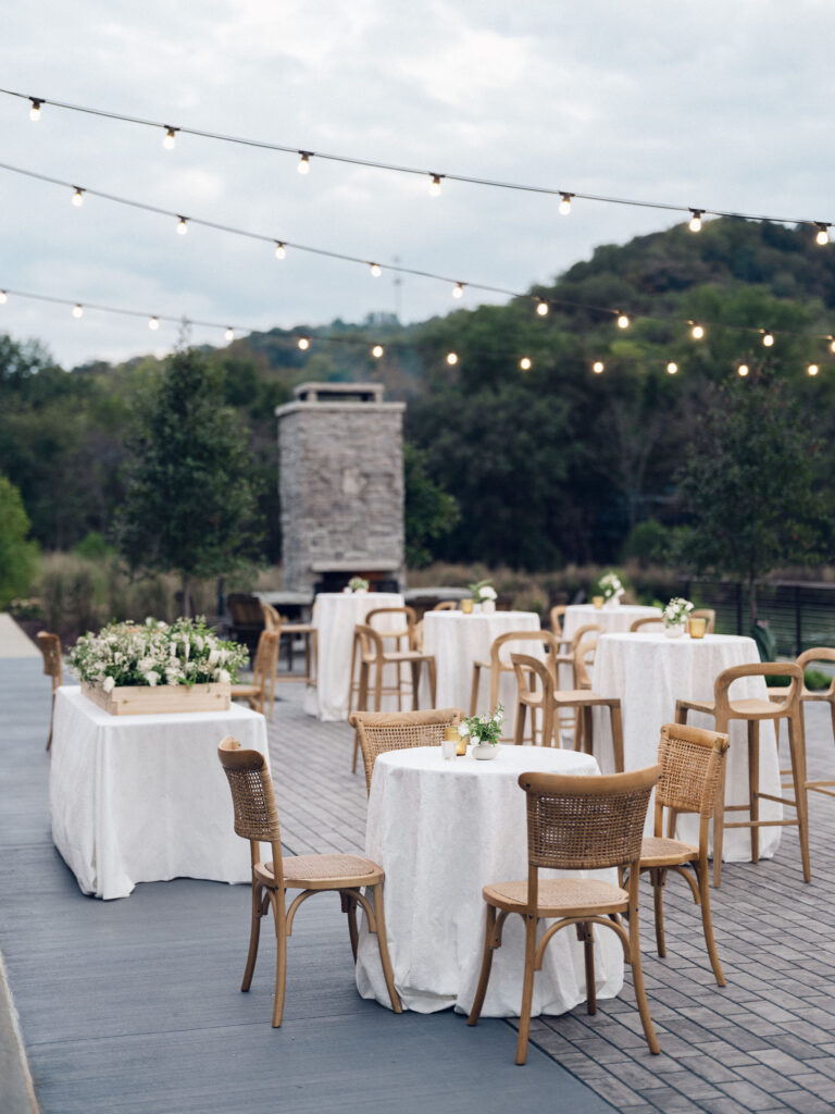 Elegant outdoor cocktail hour with bud vase floral arrangements scattered throughout high boy tables. All-white roses, lisianthus, delphinium, spray roses, ranunculus, campunala, saponaria, and light greenery were used for bud vases to correspond with floral reception centerpieces. The escort display was highlighted with chamomile, flannel flower, ranunculus, cloud garden roses, spray roses and greenery. Design by Rosemary and Finch Floral Design in Nashville, TN. 